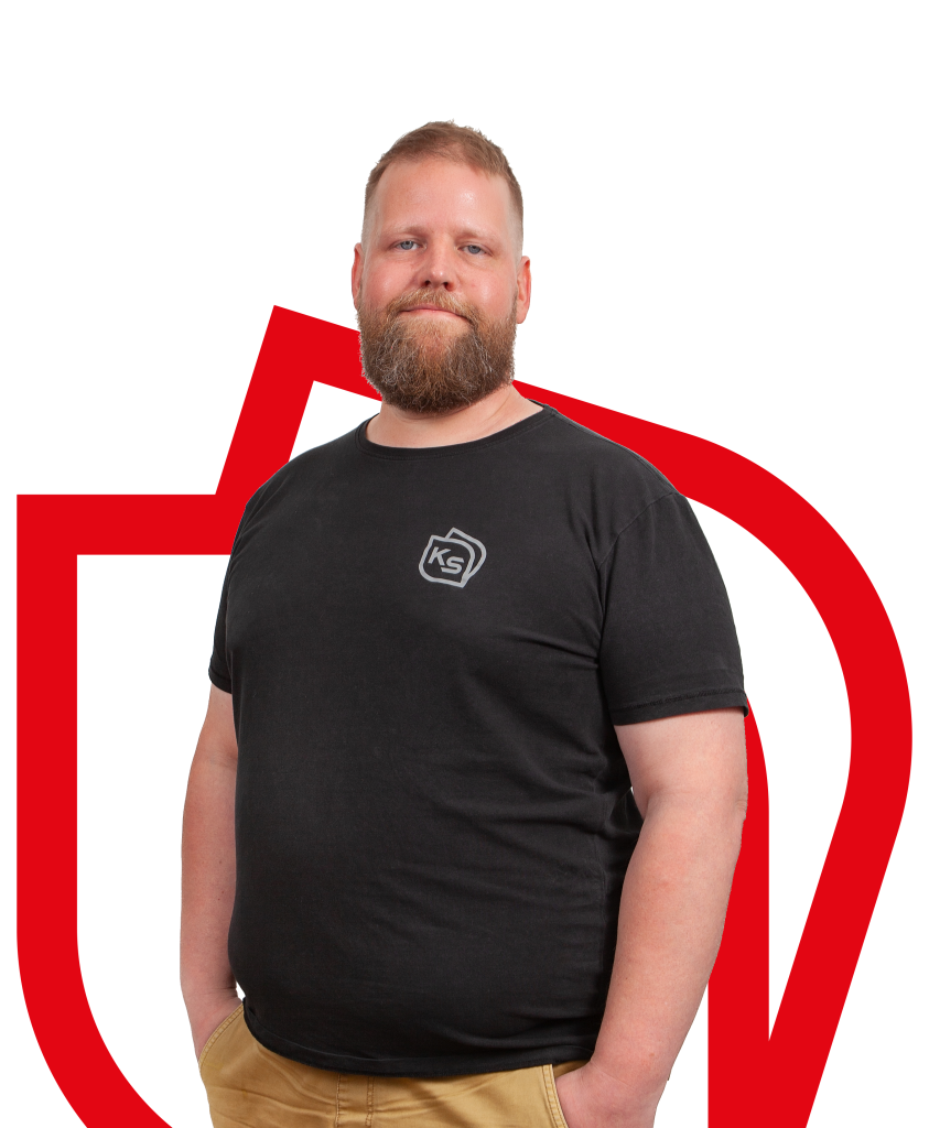 Mieshenkilö seisoo kädet taskussa, musta lyhythihainen trikoopaita päällä, katsoo suoraan kohti ja hymyilee.